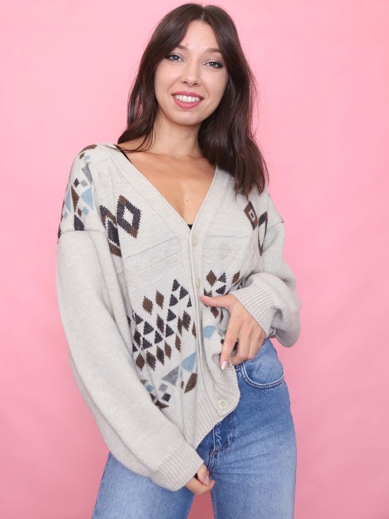 Model wearing a neutral-toned vintage cardigan with Aztec geometric knit design, styled with high-waisted jeans against a pink background.