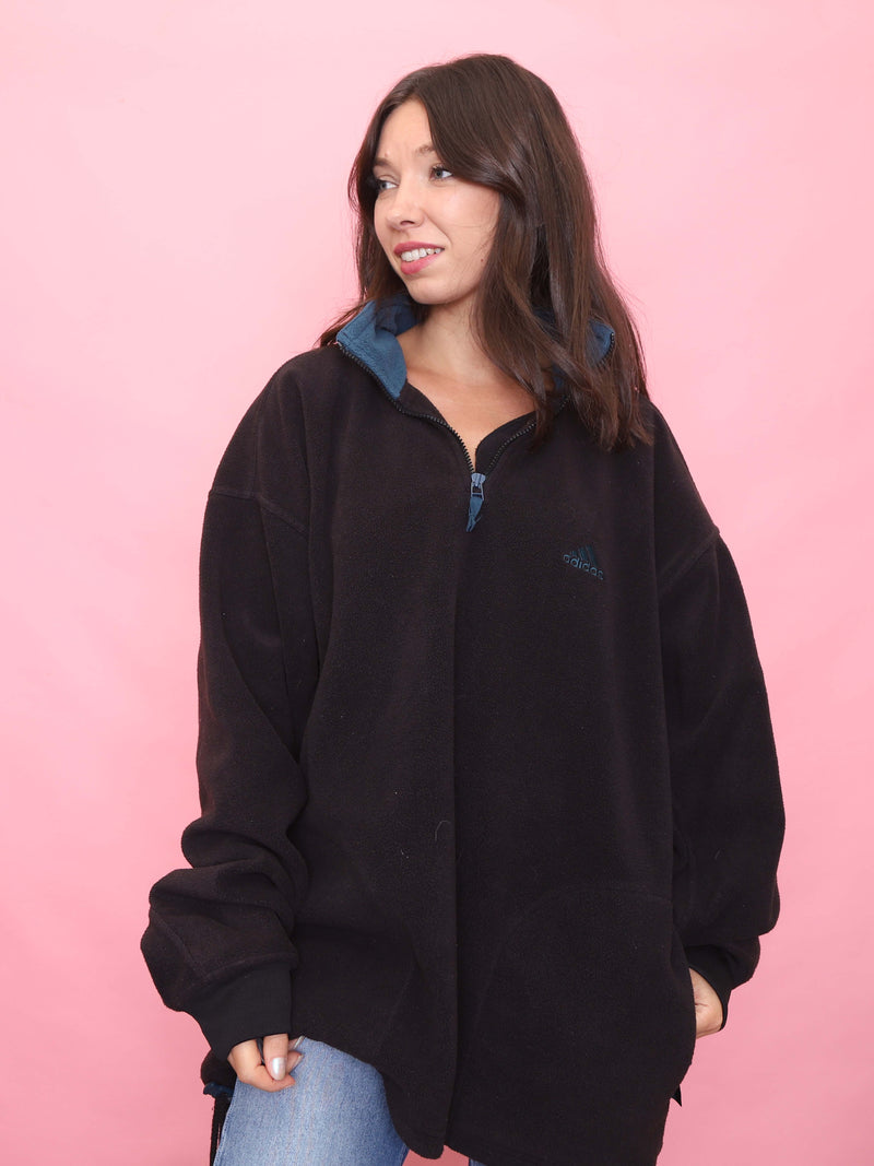 Model wearing a vintage black Adidas fleece with 1/4 zip front and teal collar against a pink background.